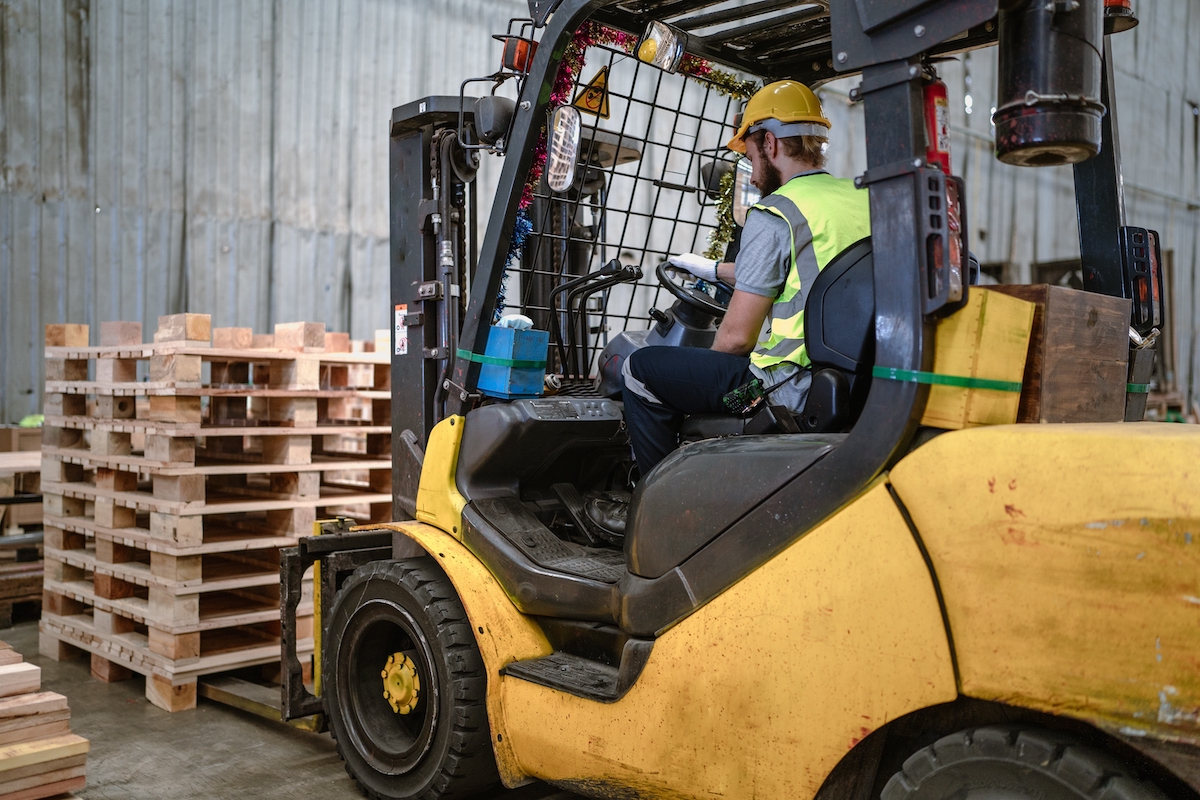 Yellow forklift truck operator wearing hard hat and hi-vis vest transporting wooden pallets in industrial warehouse